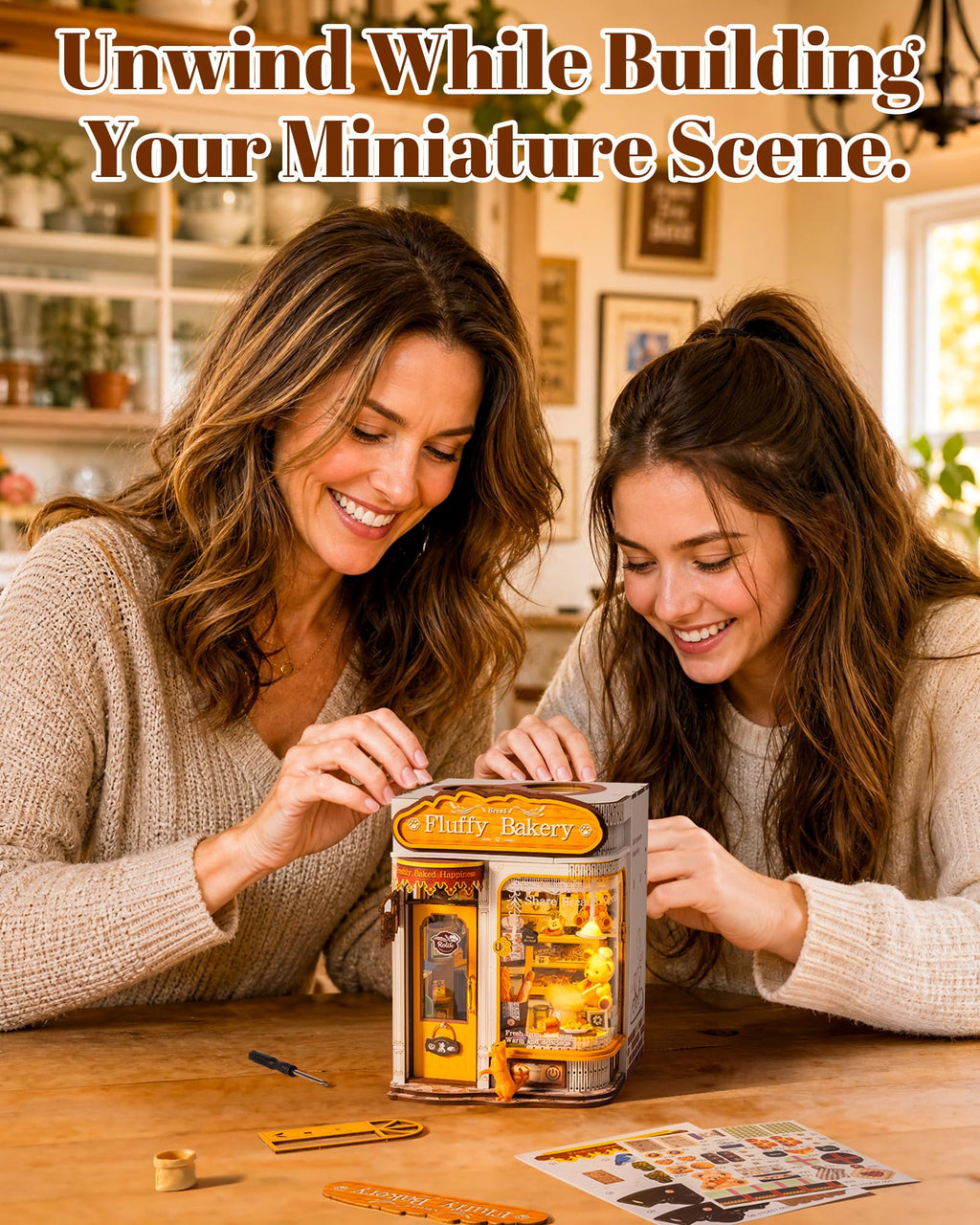 Two women smiling and working together at a wooden table with a miniature dollhouse scene labeled 'Bakery' in the center. Text at the top reads 'Unwind While Building Your Miniature Scene.