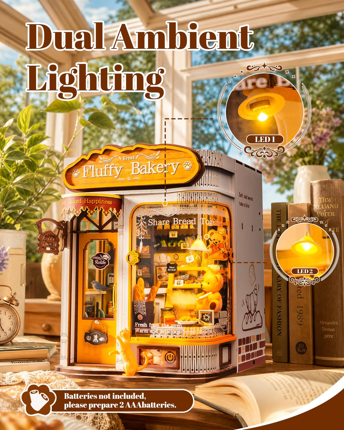 A miniature LED lighted bakery model displayed on a tabletop with books and plants in a sunlit room. Large text reads 'Dual Ambient Lighting,' with two circular inset close-ups showing warm yellow LED glow inside the bakery.