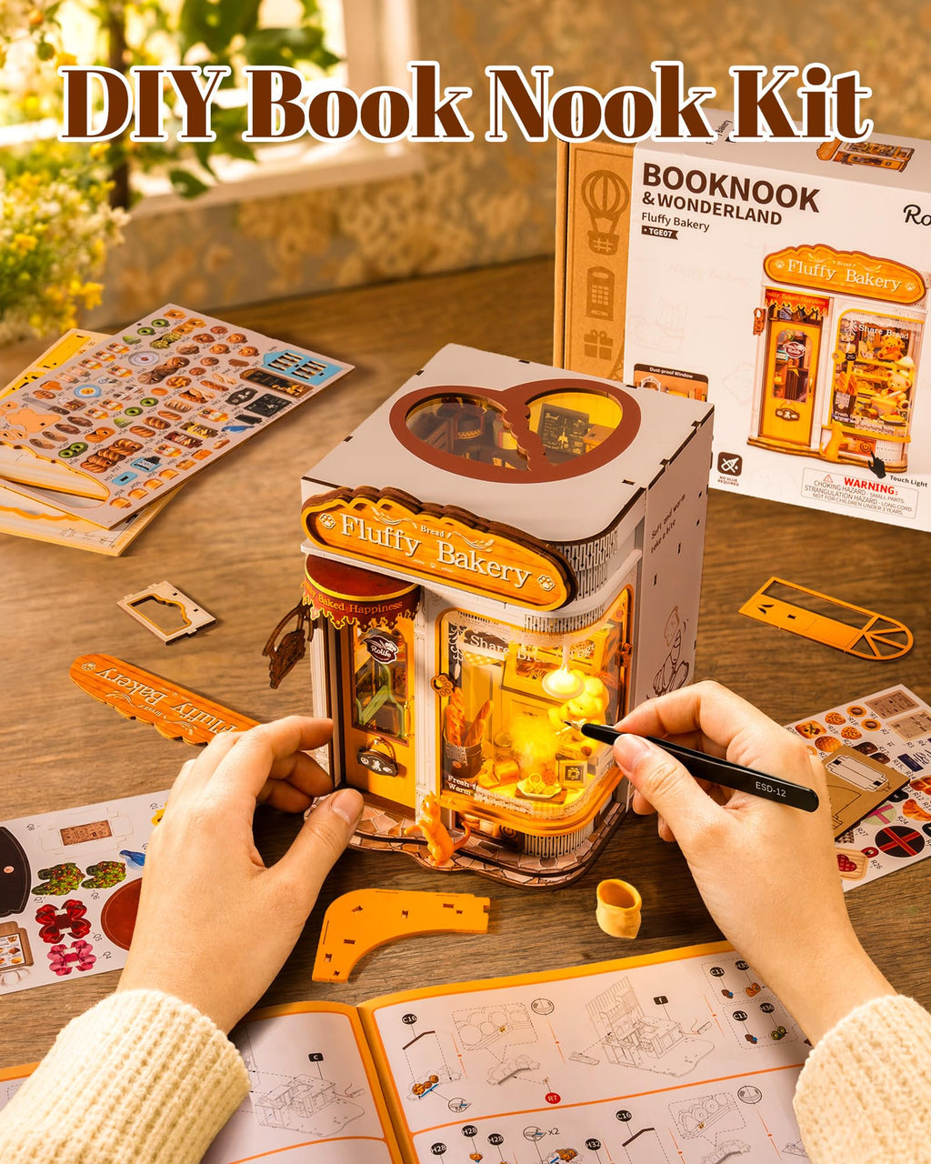 DIY Book Nook Kit showing a completed 3D wooden bookshelf model styled as a bakery, with hands assembling or decorating it. The scene includes the product box, sticker sheets, and instruction pages on a wooden table, with 'DIY Book Nook Kit' text overlay at the top.