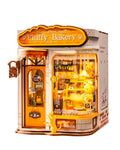 A 3D wooden model kit of a bakery storefront named 'Fluffy Bakery', featuring a detailed door, window display with plush toys, and decorative elements, shown on a solid white background.