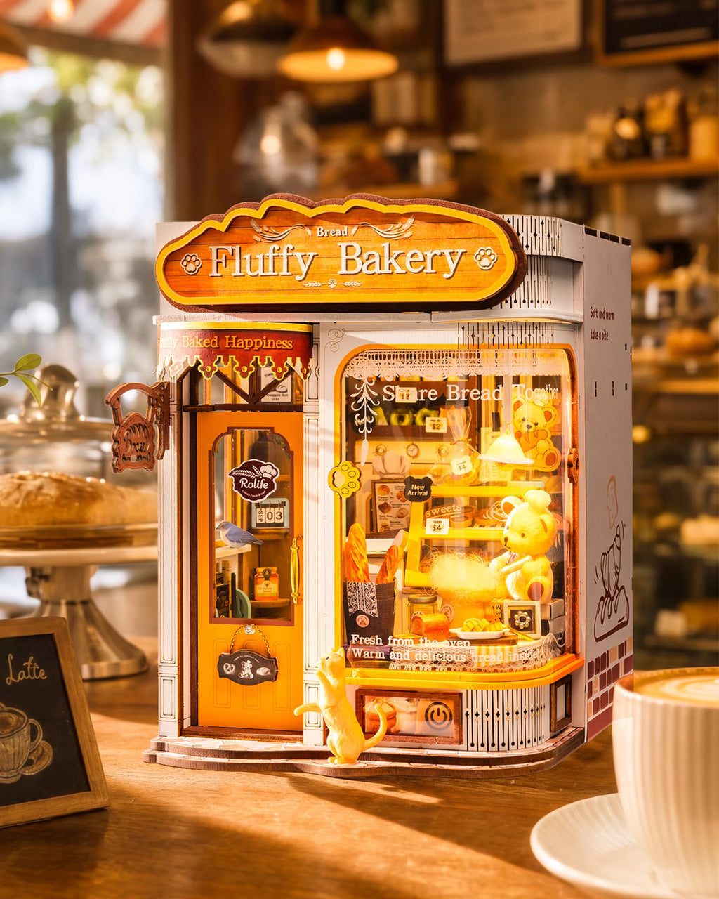 A miniature Fluffy Bakery model displayed on a wooden table in a cozy, warm-toned bakery setting. The white and orange bakery structure features a glowing yellow interior with tiny shelves, pastries, and a small plush character, with a sign reading 'Fluffy Bakery' on top.