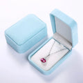 A silver pendant necklace with a heart-shaped pink gemstone is displayed inside an open light blue velvet jewelry box next to its closed lid on a white background.