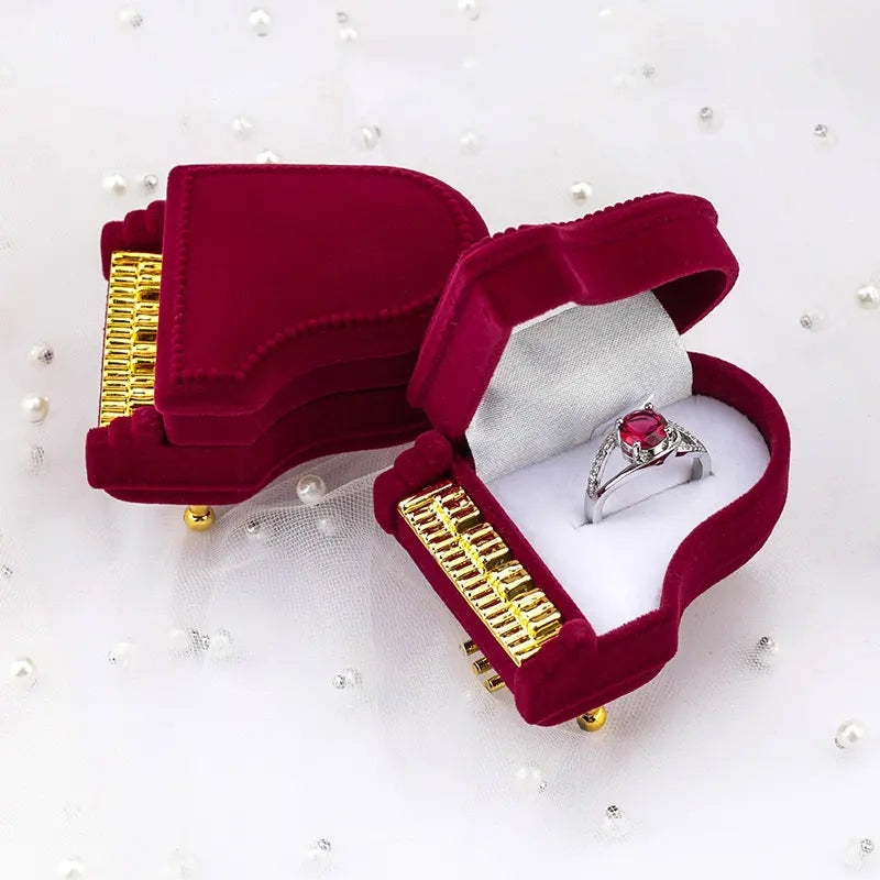Open red velvet piano jewelry box displaying a ruby and diamond ring in white satin interior surrounded by scattered pearls on white background