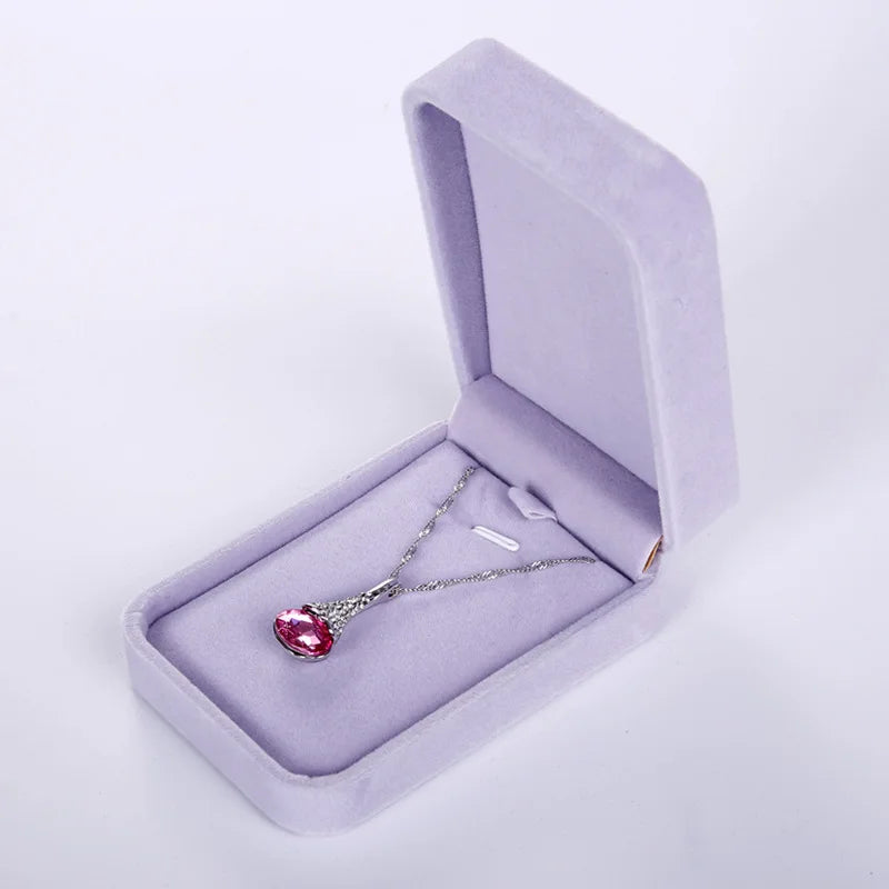 A silver pendant necklace with a heart-shaped pink gemstone, displayed inside an open light purple velvet jewelry box on a white background.