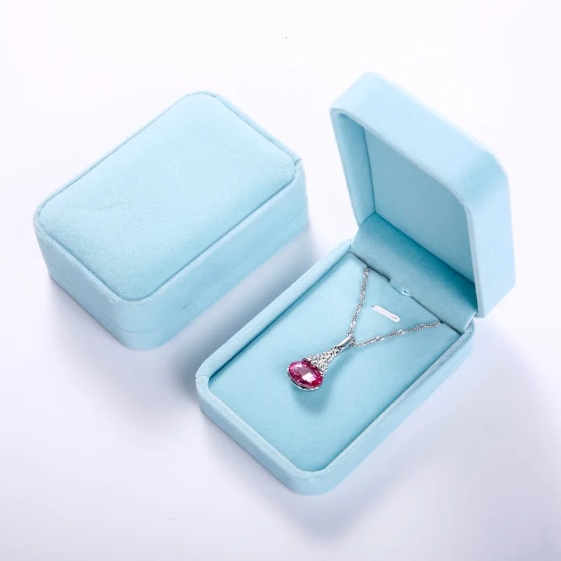A silver pendant necklace with a pink heart-shaped gemstone displayed inside an open light blue velvet jewelry box, with a second closed matching box beside it on a white background.