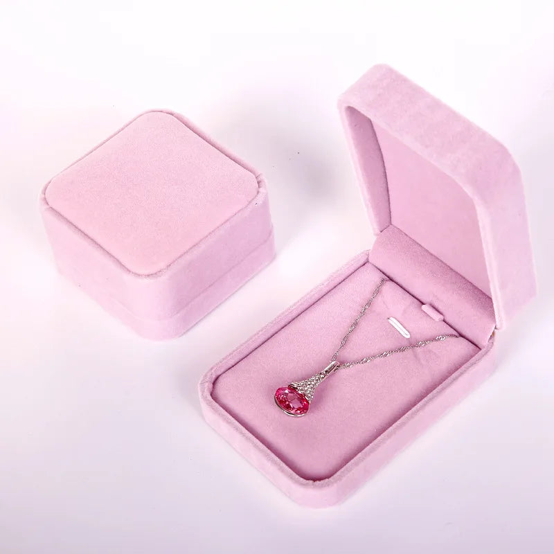 A pink velvet jewelry box shown open with a silver necklace featuring a pink heart-shaped pendant inside, alongside a closed matching pink box on a white background.