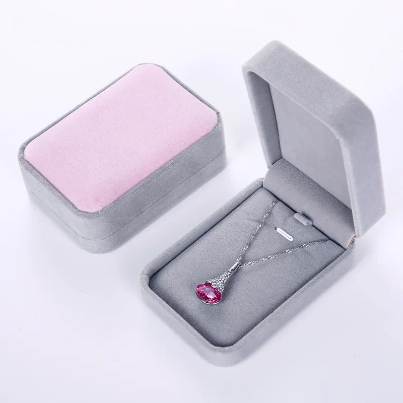 A silver pendant necklace with a heart-shaped pink gemstone is displayed inside an open gray velvet jewelry box, with a closed matching box shown beside it on a white background.