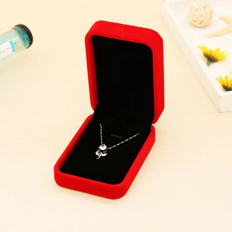 A silver pendant necklace displayed inside an open red velvet jewelry box with a black interior, set on a light beige surface with decorative props in the background.