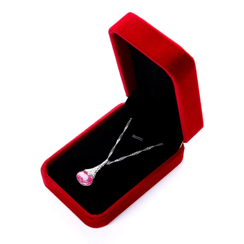 A silver-toned pendant necklace with a teardrop-shaped pink gemstone, displayed inside an open red velvet jewelry box on a white background.