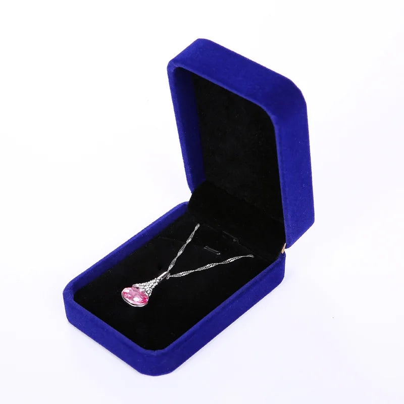 A silver necklace with a heart-shaped pink gemstone pendant, displayed inside an open royal blue velvet jewelry box on a white background.