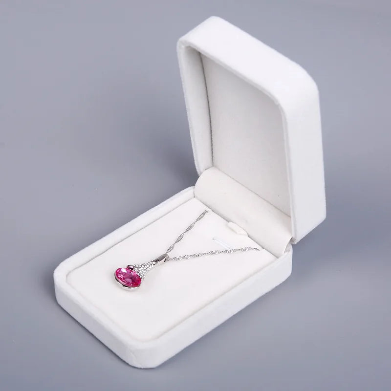 A silver pendant necklace with a heart-shaped pink gemstone, displayed inside an open white jewelry box on a neutral gray background.